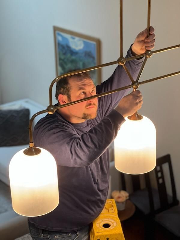 A person on a yellow ladder reaching up to adjust or install a brass chandelier with two glowing white glass shades.