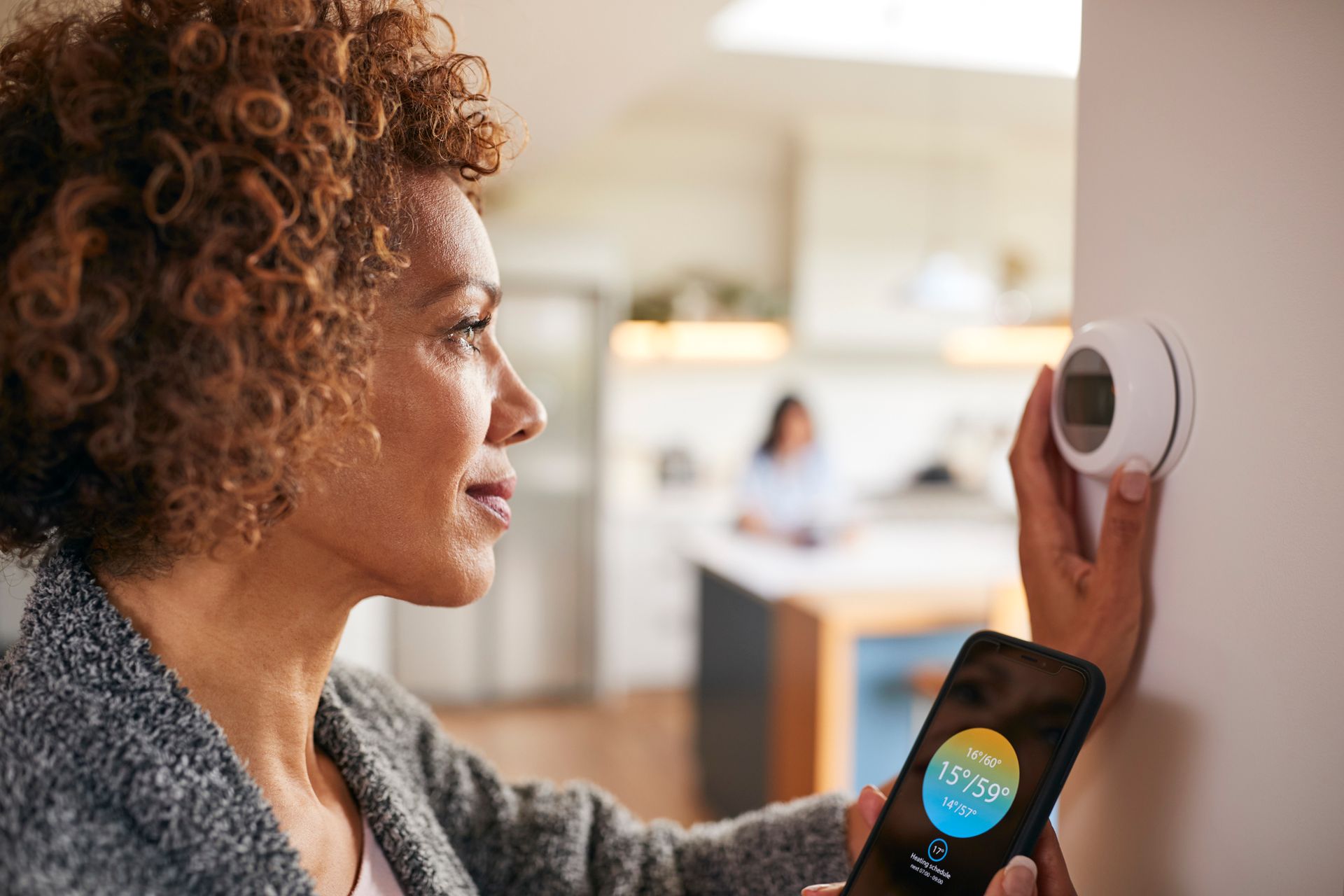 A person adjusting a wall-mounted smart thermostat while holding a smartphone displaying the thermostat app.