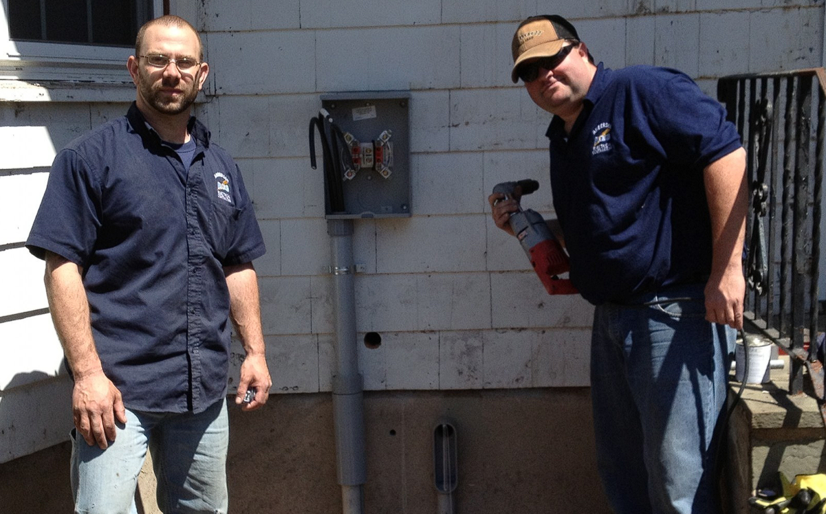 Two workers in blue uniforms stand outside a building next to an open electrical meter box; one holds a power tool.