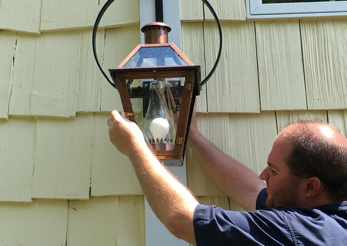 A person adjusts a copper-framed outdoor lantern mounted on a light yellow shingled wall.