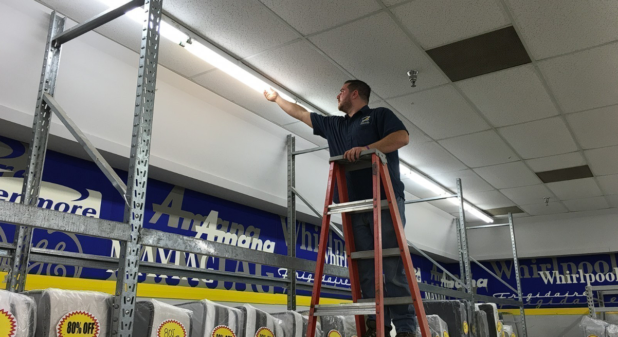 A person stands on a red stepladder, reaching up to adjust a long fluorescent light fixture in a retail warehouse store.