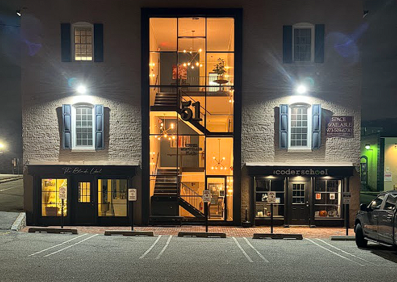 A three-story brick building at night with a central glass tower showing a staircase, flanked by lit retail storefronts.