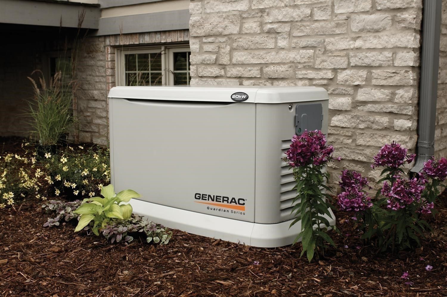 A grey Generac standby generator installed on a landscaped garden bed next to a stone house wall.