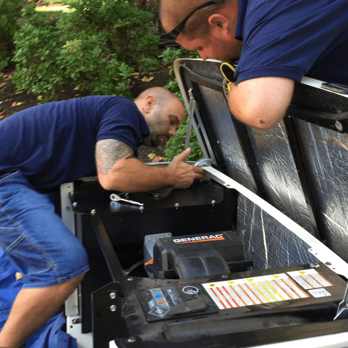 Two individuals in dark blue shirts inspect the open motor compartment of a generator outdoors.