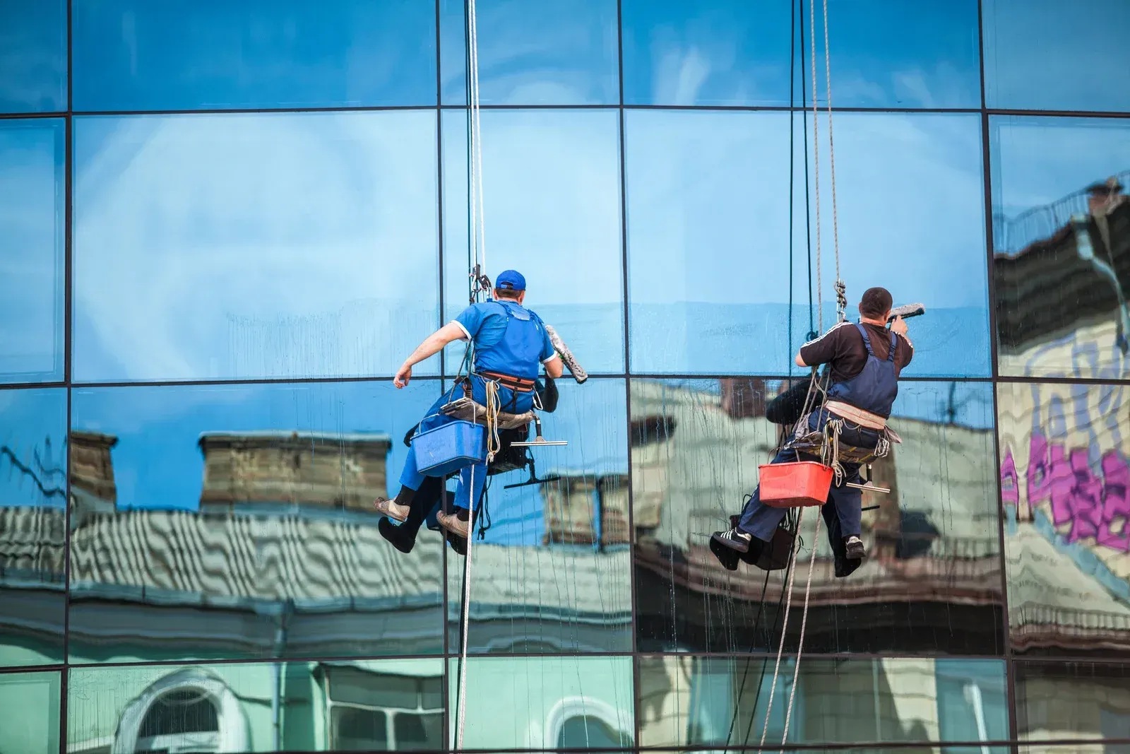 Two window washers suspended on ropes, cleaning a glass building; reflected buildings and sky.