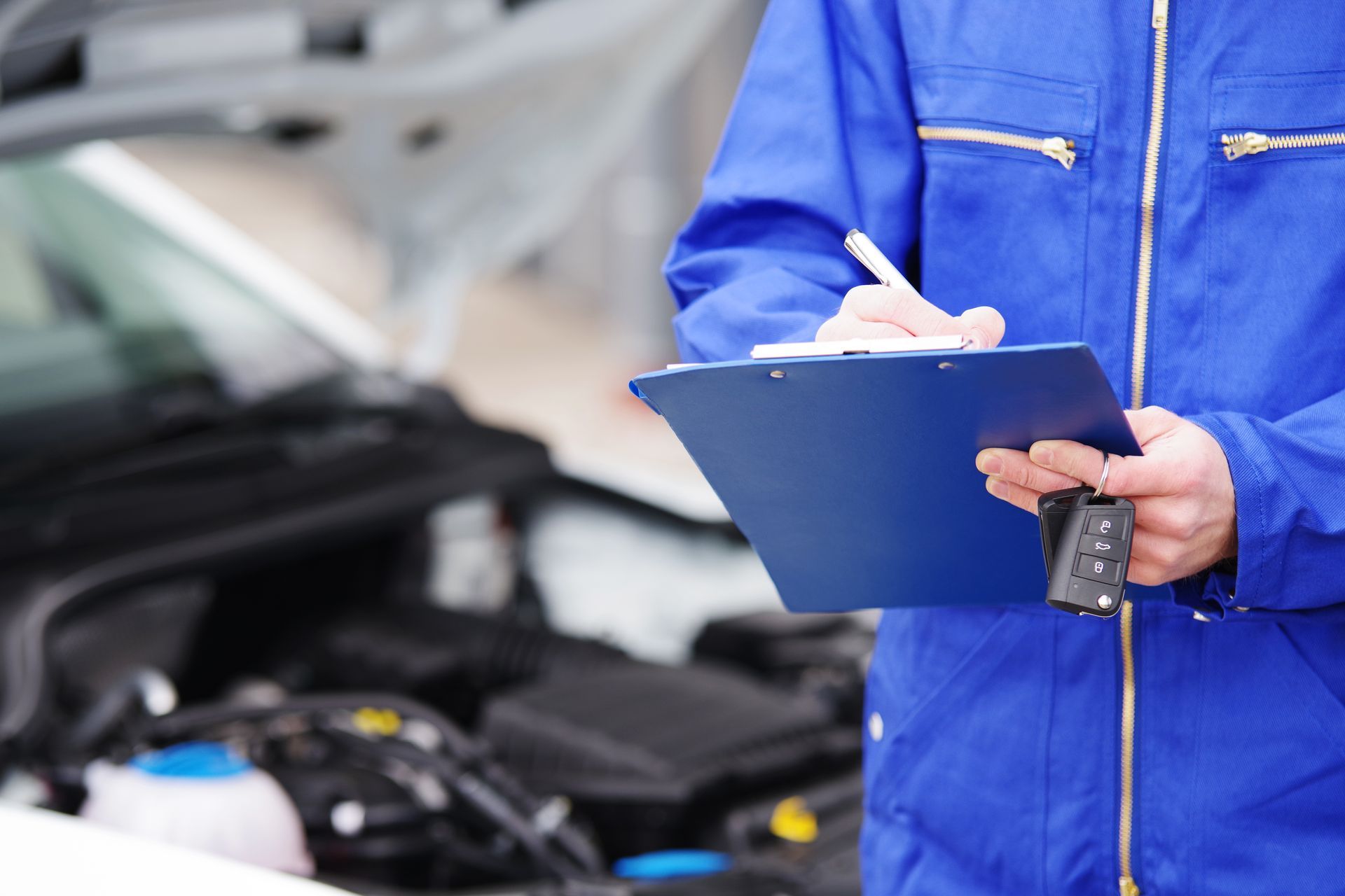 Mechaniker im blauen Overall schreibt auf einem Klemmbrett, im Hintergrund der Automotor.