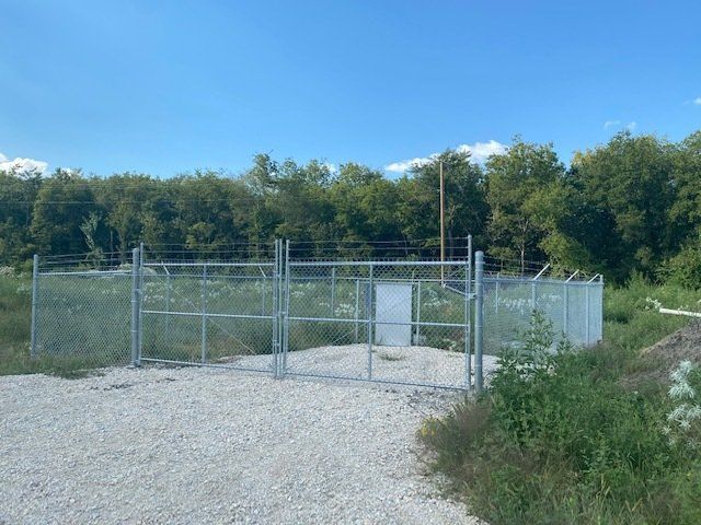 A metal fence surrounds a gravel area with trees in the background.