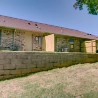 Stone building with a sloped roof and small windows behind a low retaining wall and grassy yard
