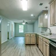 Empty modern kitchen with light wood cabinets, island, and pale blue walls, viewed toward a windowed dining area