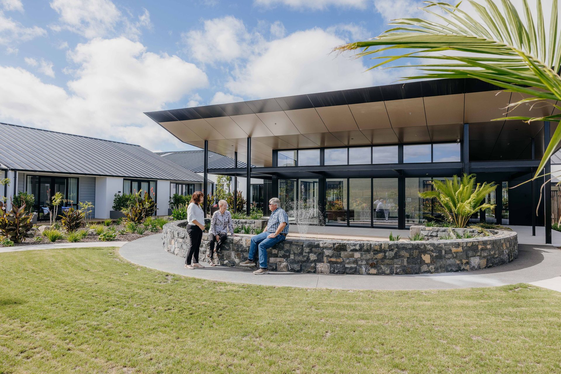 People sit by a stone wall in front of a modern home with glass windows and a curved roof.