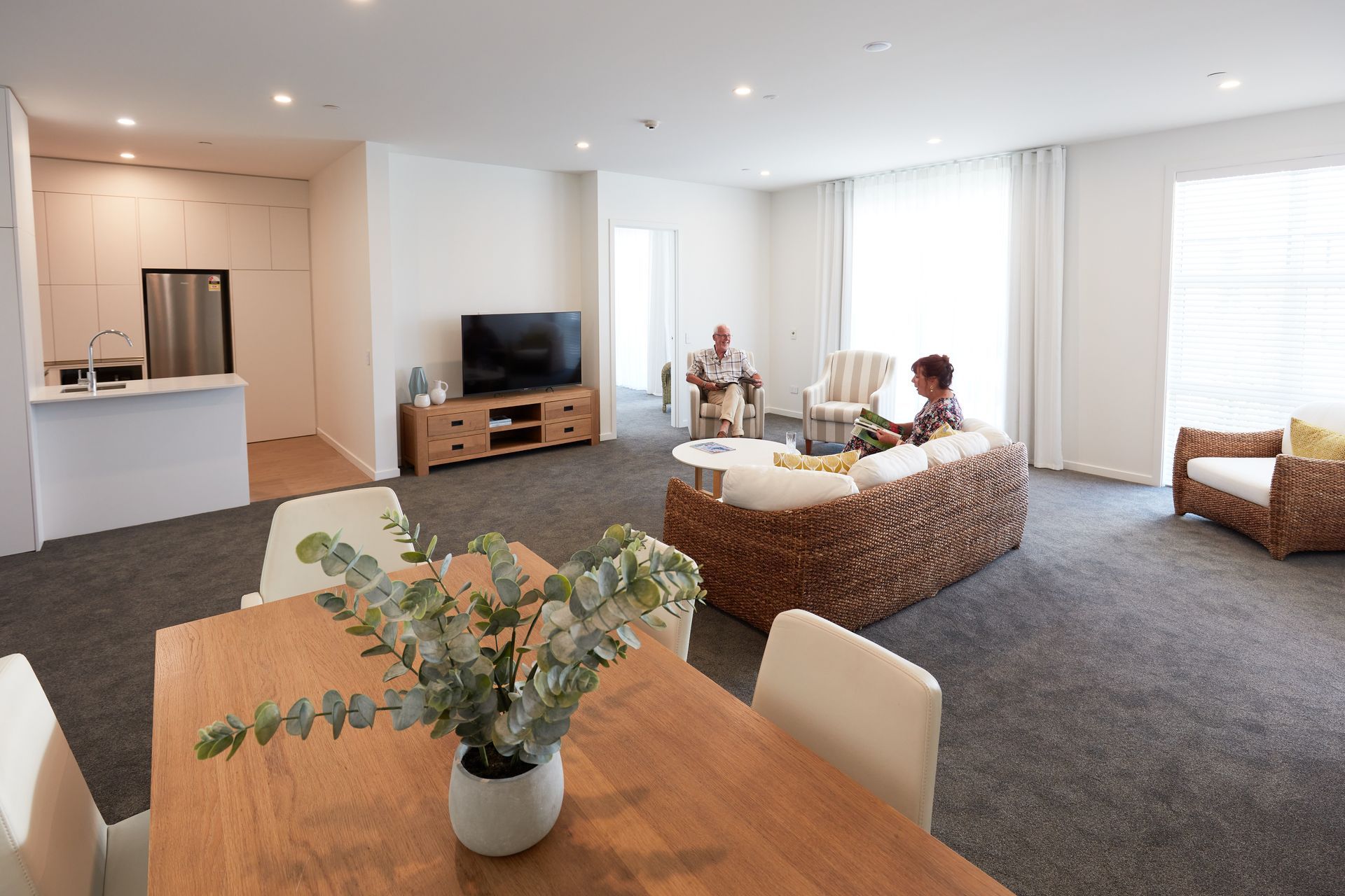 Spacious living room with seniors relaxing, featuring a dining table, seating, and a kitchenette.