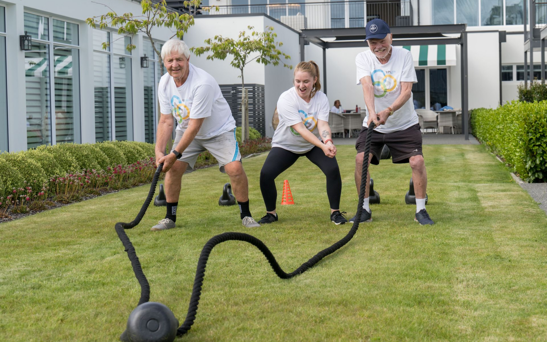 Three people exercising outdoors with battle ropes on a grassy lawn.