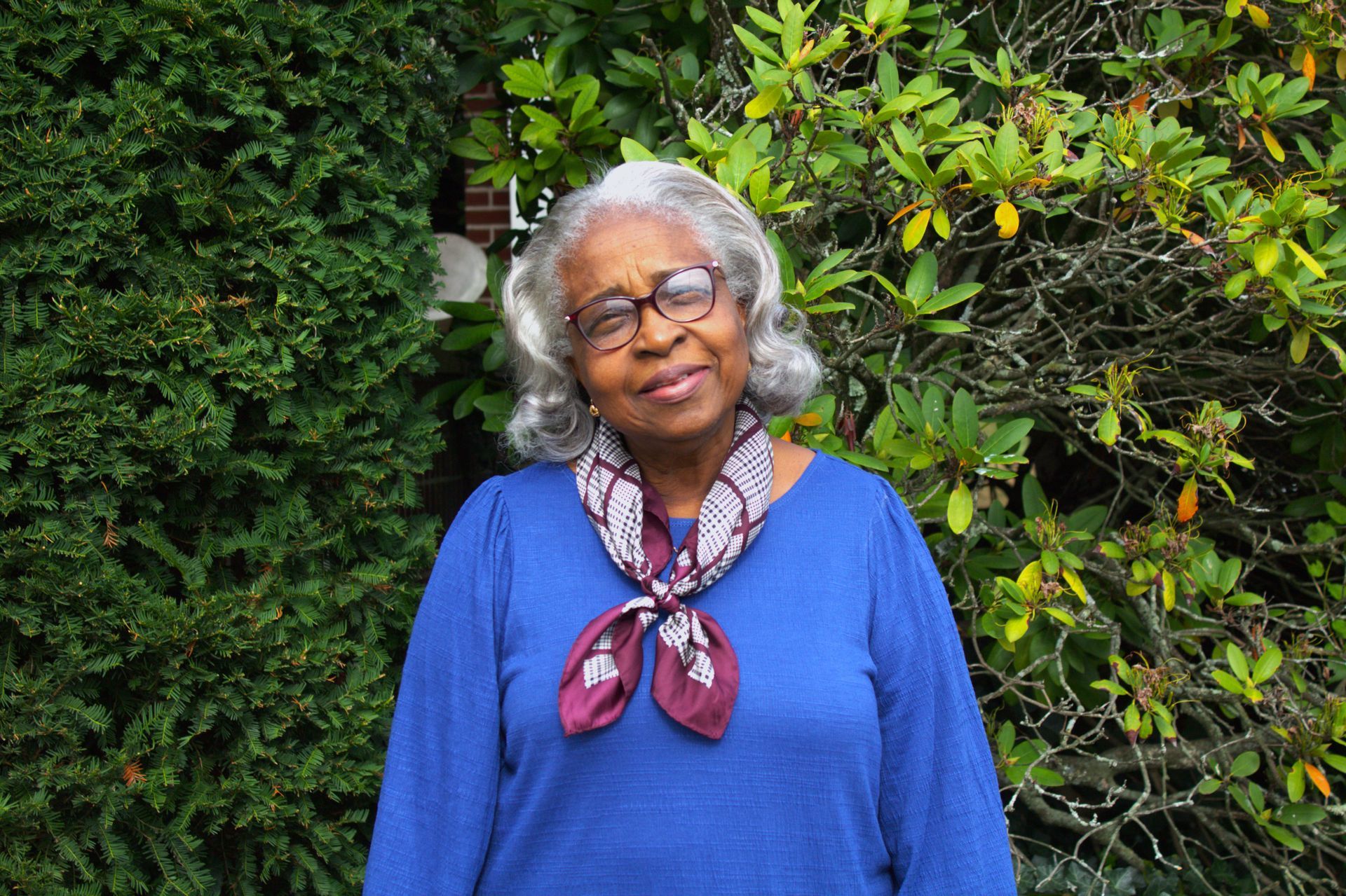 An elderly woman wearing glasses and a scarf is standing in front of a bush.