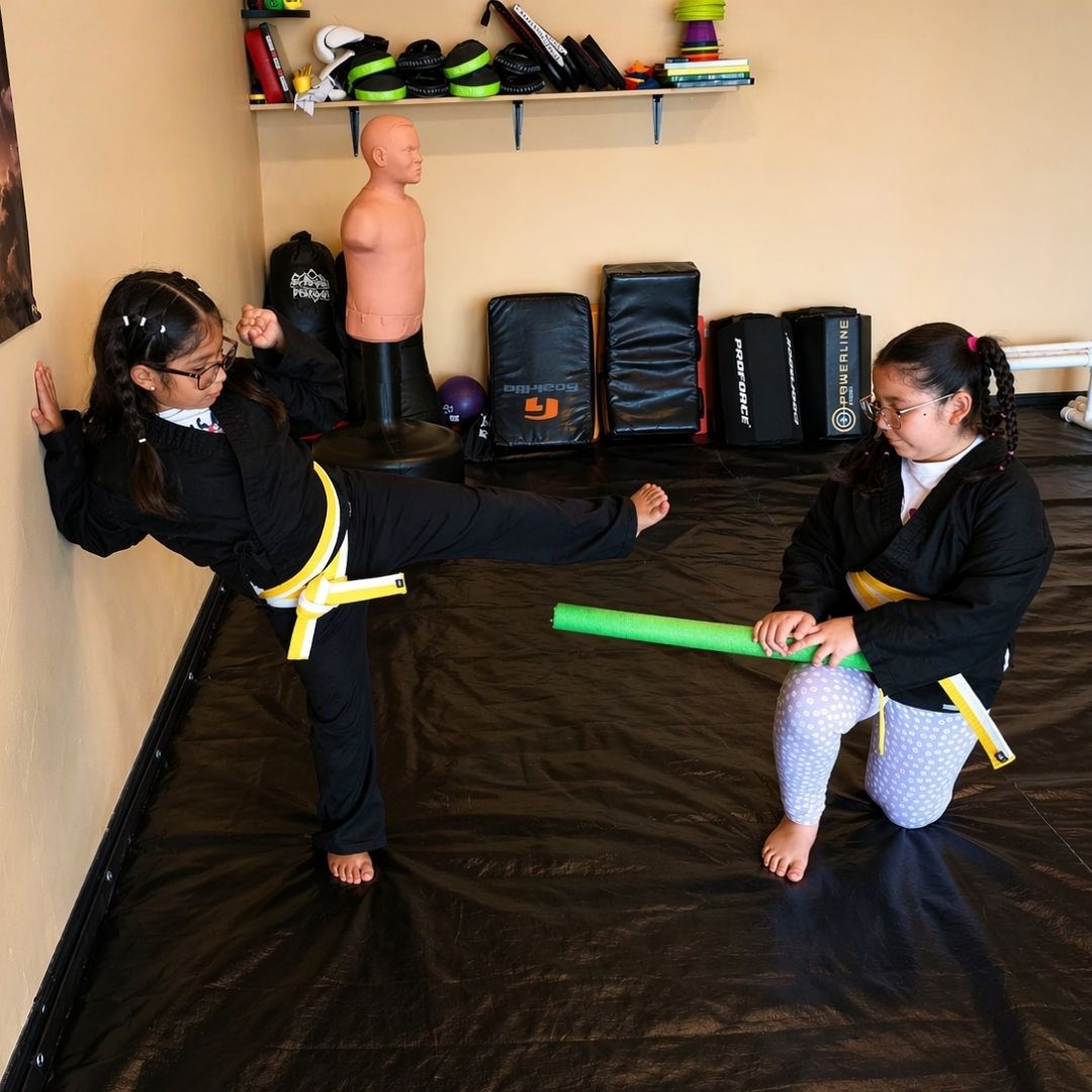 Two children practicing Brazilian Jiu-Jitsu on a mat. One child in white gi has the other in a leg lock. A girl watches.