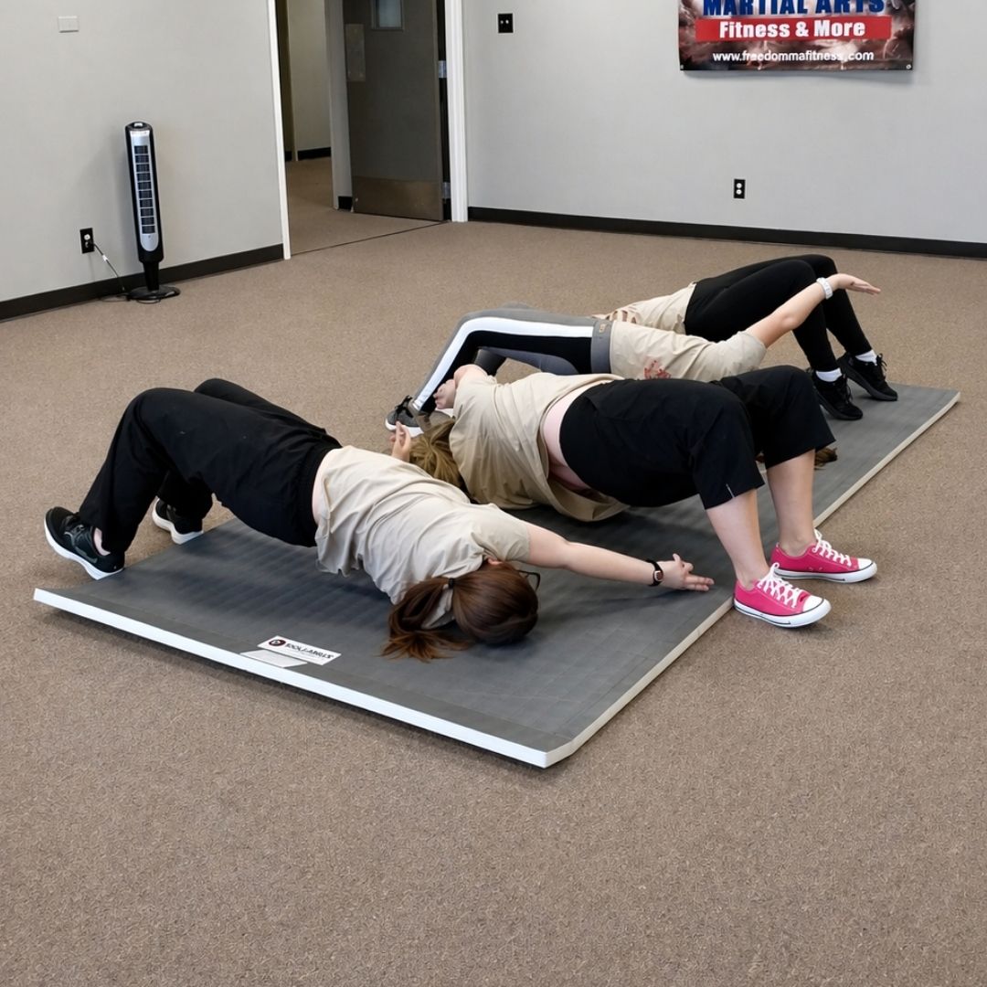 Two men in grappling position on a mat. One embraces the other from behind, both smiling. Gym setting.
