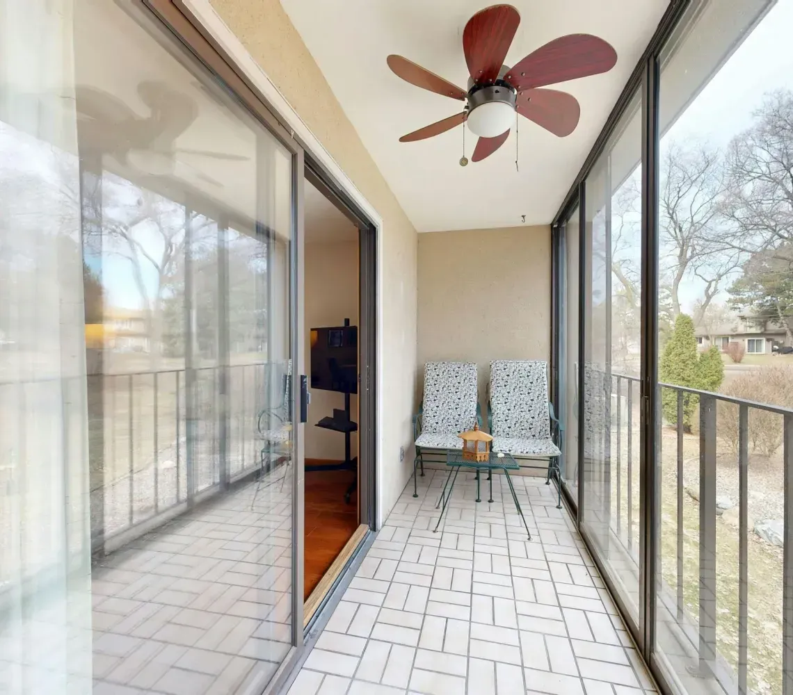 Long balcony with glass sliding doors, tiled floor, ceiling fan, and two chairs overlooking trees