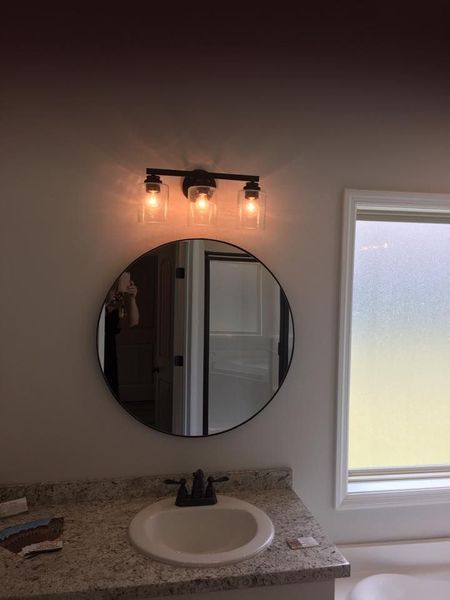 Bathroom vanity with round mirror, lit three-bulb sconce, and frosted window on the right