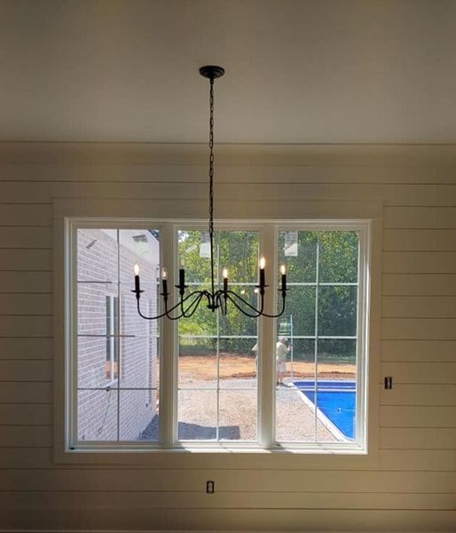 Black chandelier hanging before a large window in a light wood-paneled room, with a backyard and pool outside.