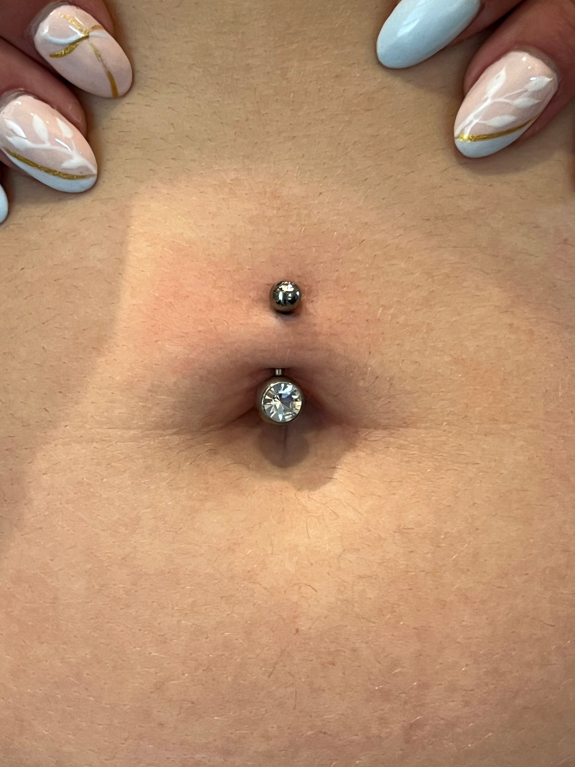 Een close-up van een navelpiercing met een zilveren staafje, een klein kraaltje bovenaan en een grotere, transparante edelsteen onderaan.