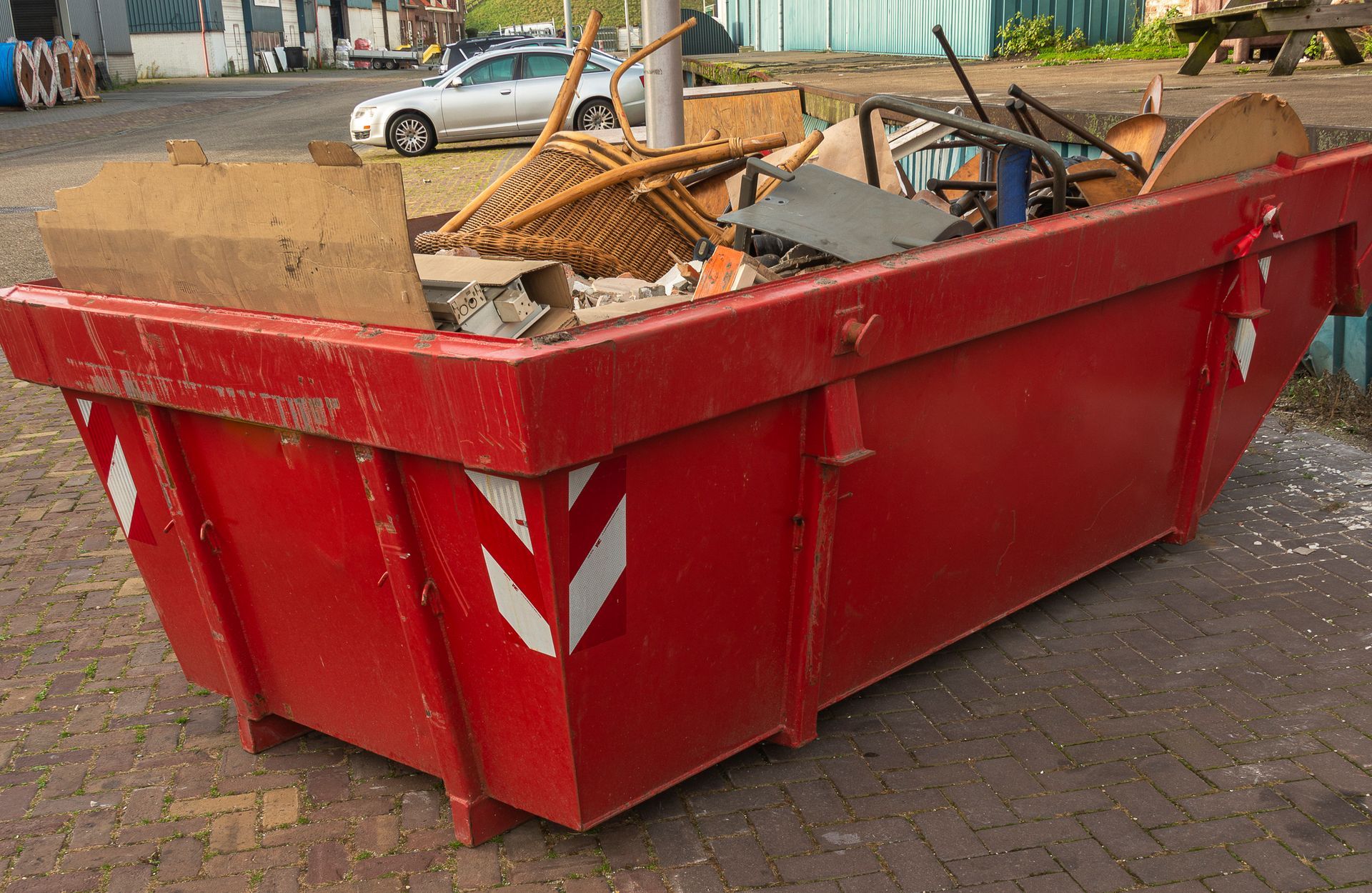 Red skip bin hire container with mixed waste, including wood and metal, placed on a paved site.