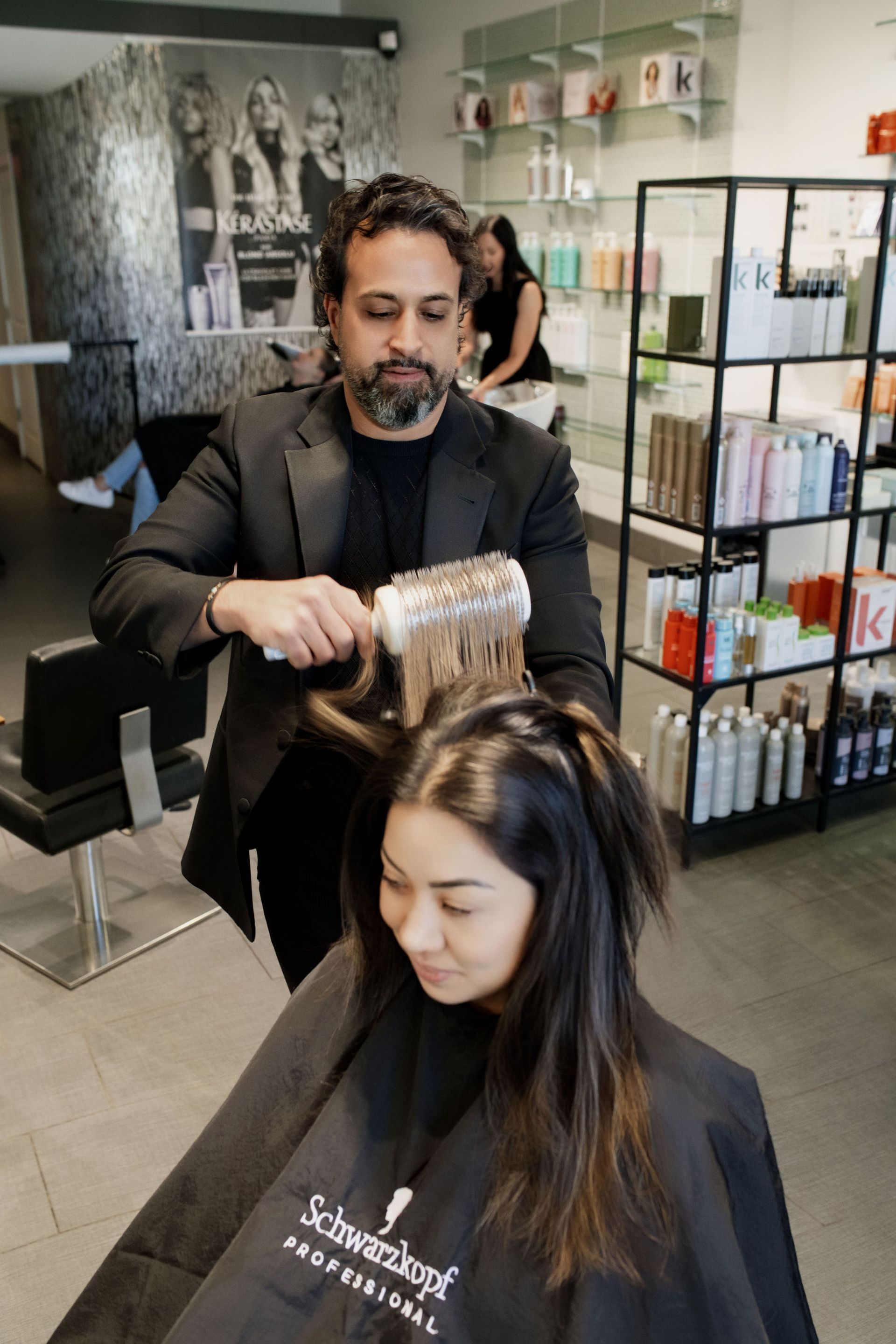 Stylist blow-drying a client’s hair in a salon, with product shelves in the background.