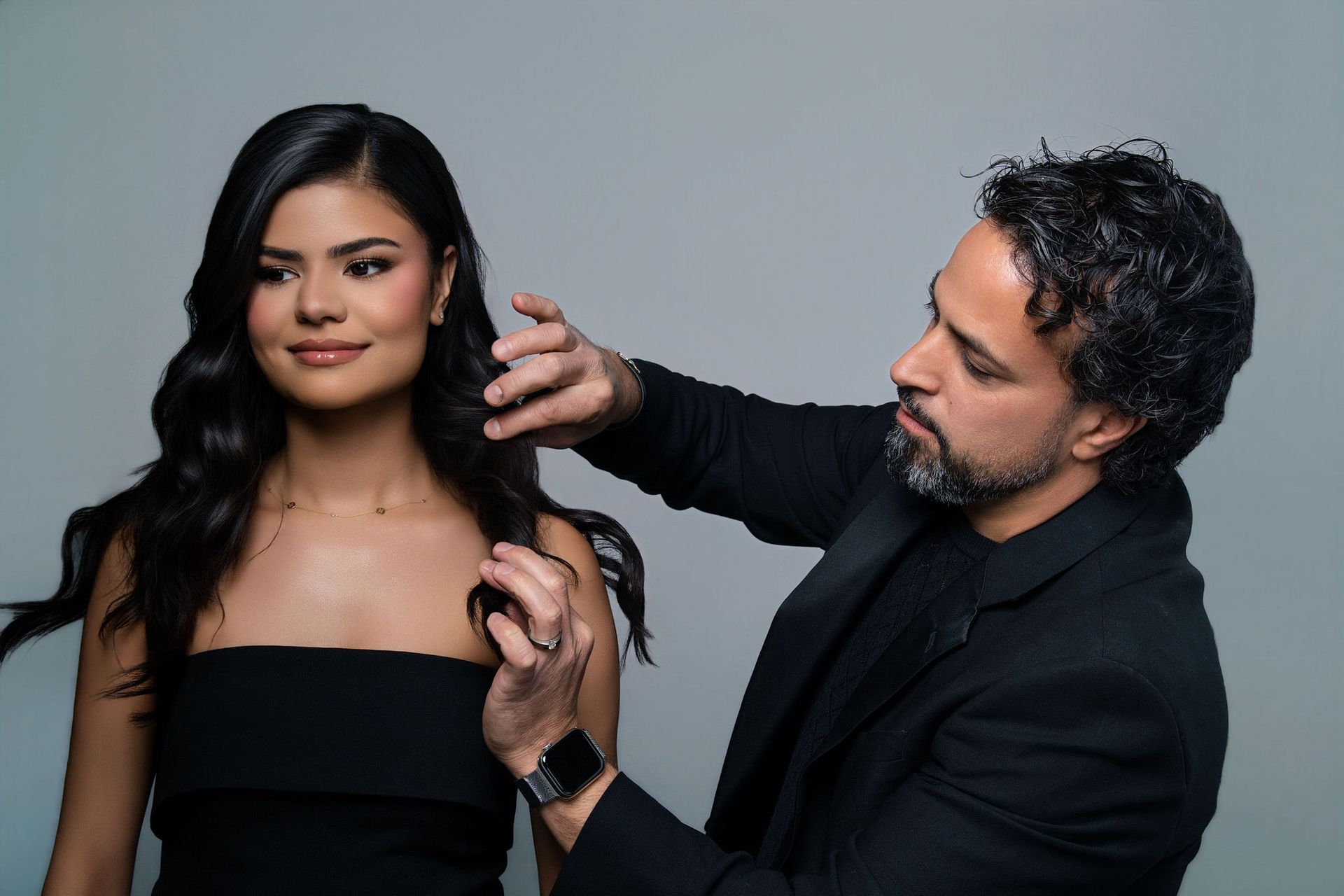 A stylist adjusting a woman's wavy dark hair. The woman wears a black top, and the background is gray.