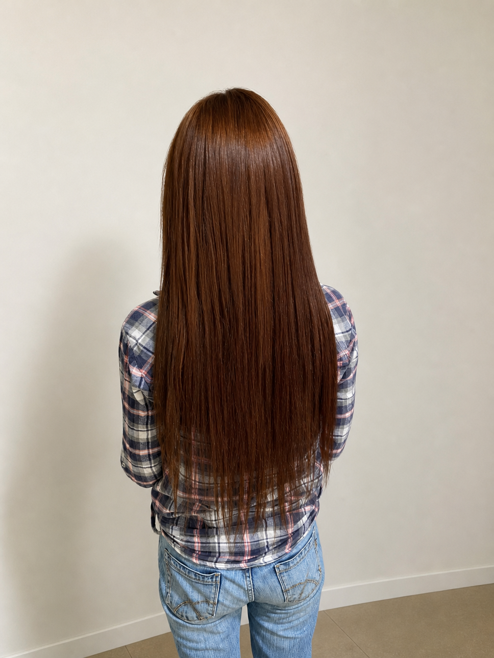 Person with long brown hair standing against a plain wall, wearing a plaid shirt and jeans — Superb Hair in Wyong, NSW