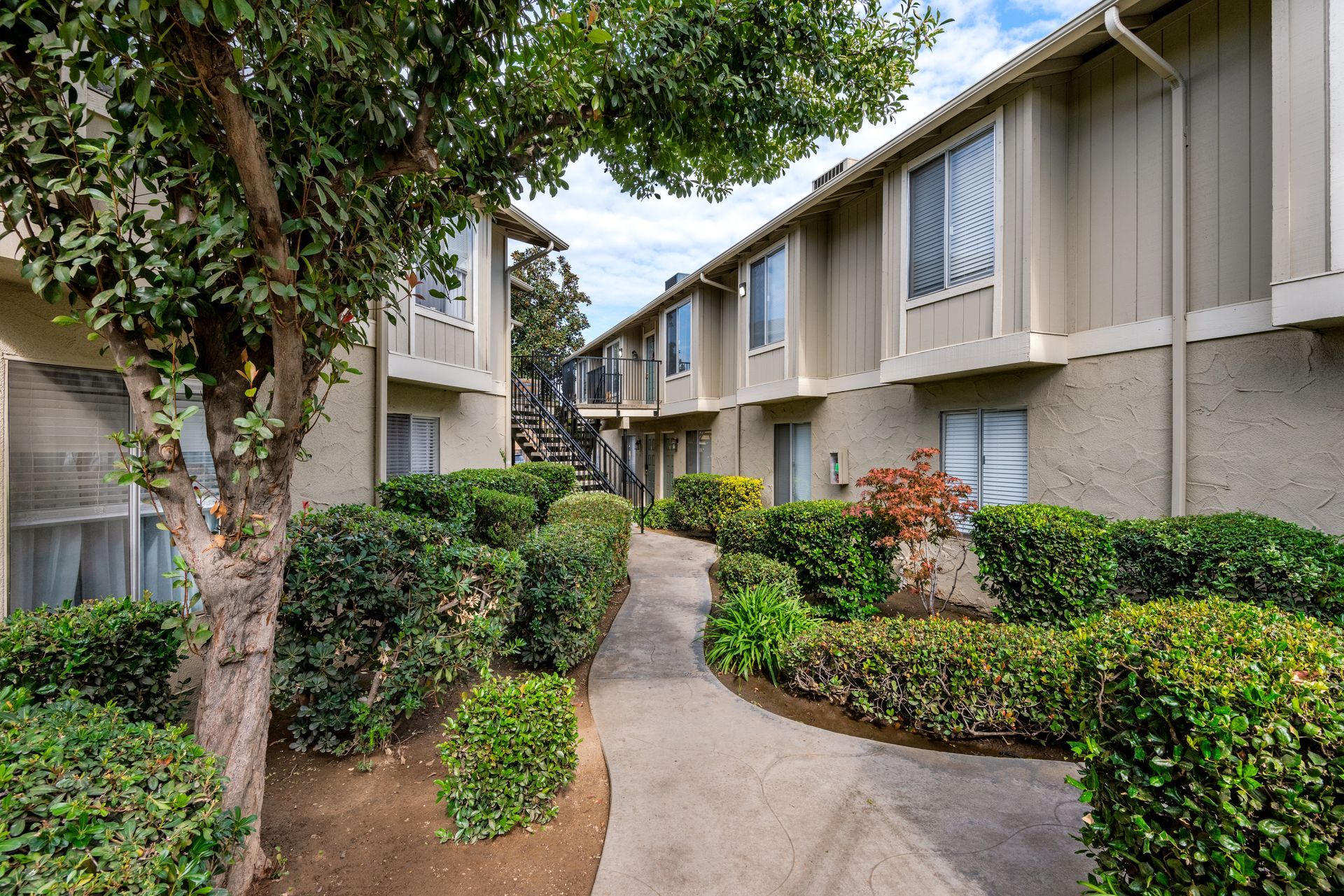 Apartment complex exterior with a path, bushes, and trees under a cloudy sky.