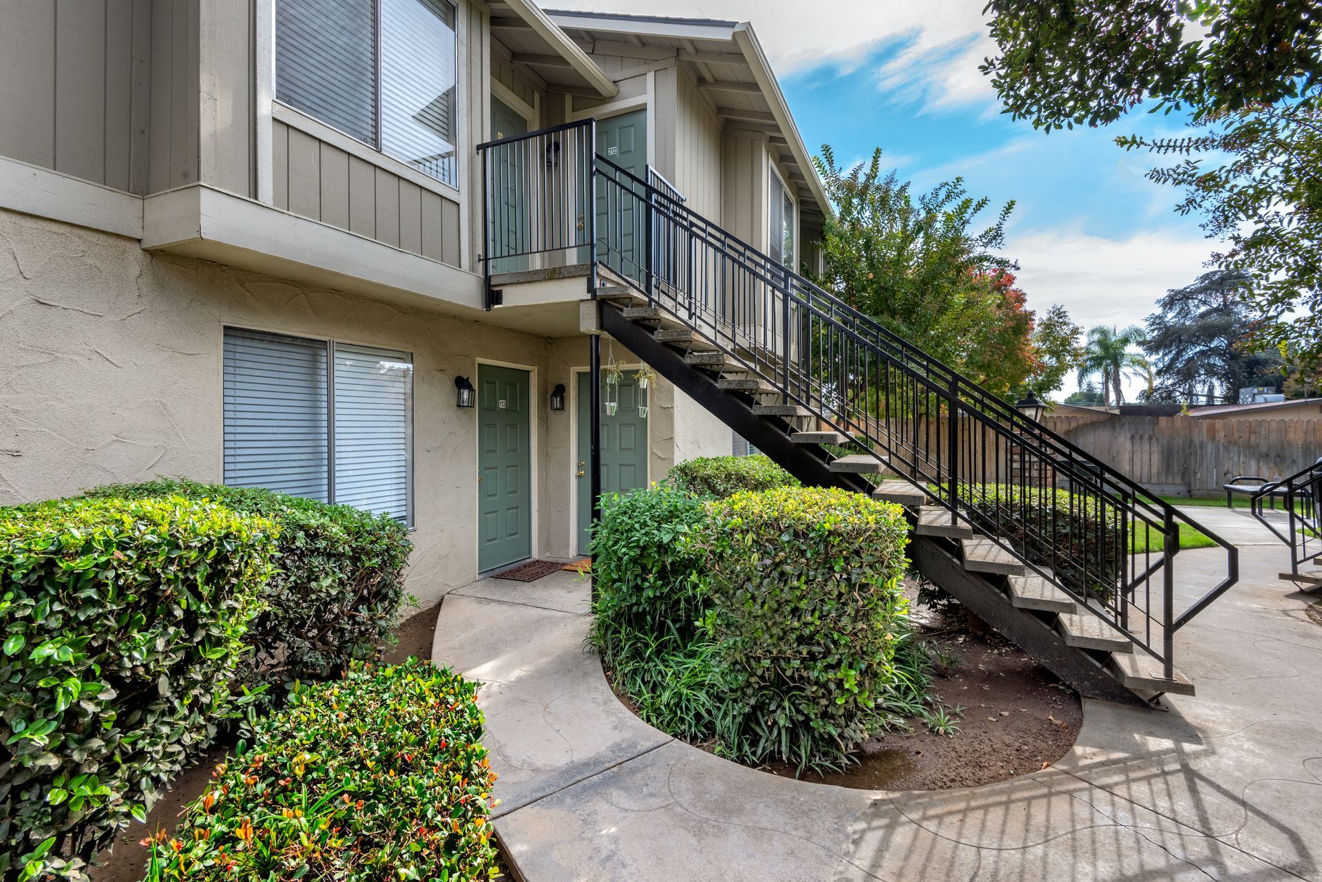 Apartment building exterior with stairs and green bushes. Concrete pathway leads to doors with green accents.