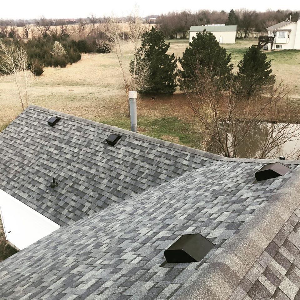 Two gray shingled roof sections with black vents. Rural landscape with trees and houses in background.