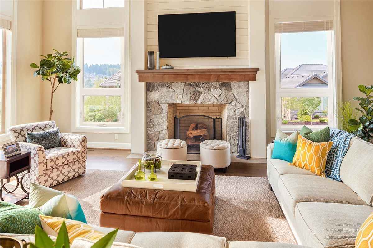 Living room with fireplace, stone surround, TV, sofa with colorful pillows, armchair, and large windows.