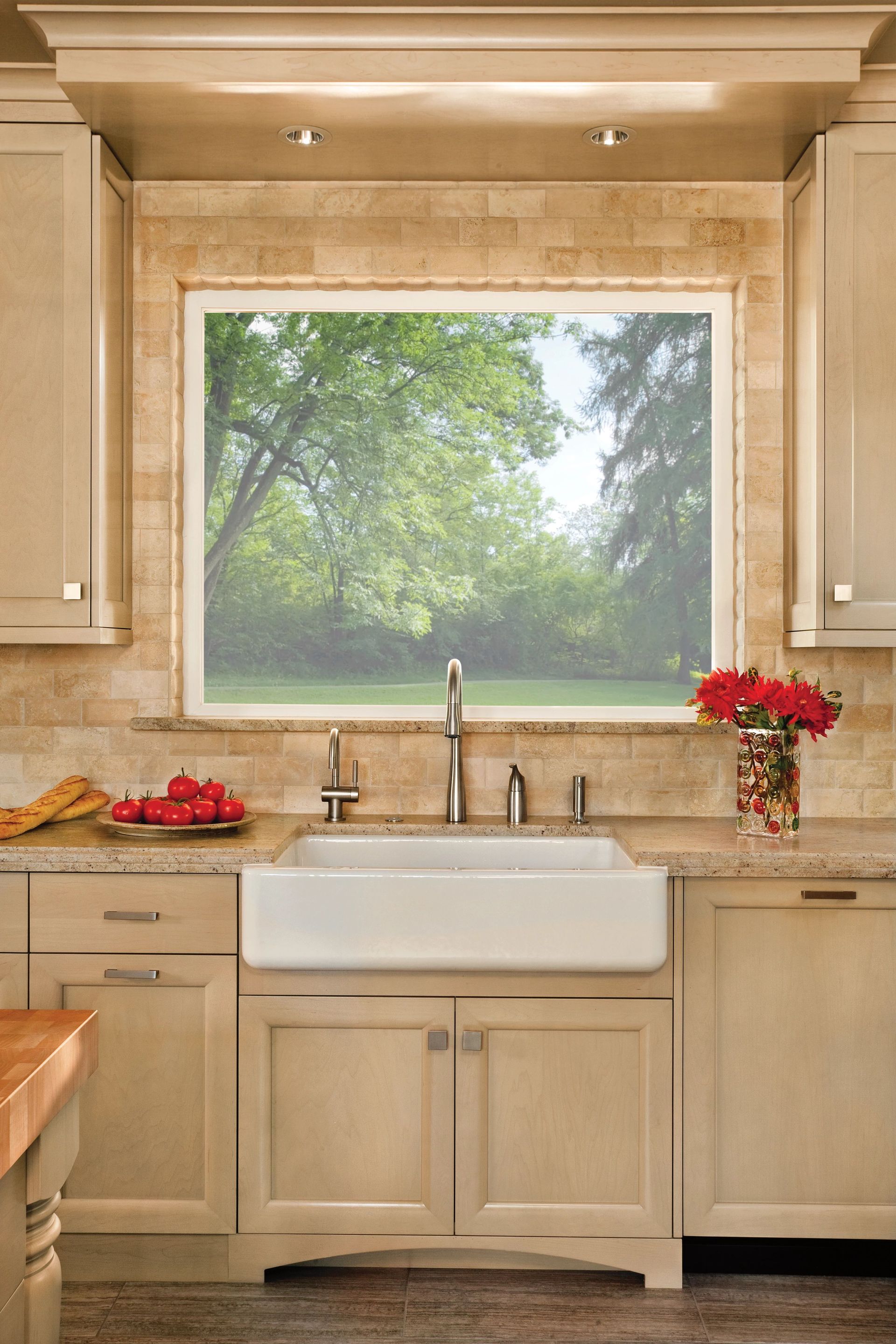 A farmhouse sink in a neutral-toned kitchen with beige cabinets, stone tile backsplash, and a large window facing trees.