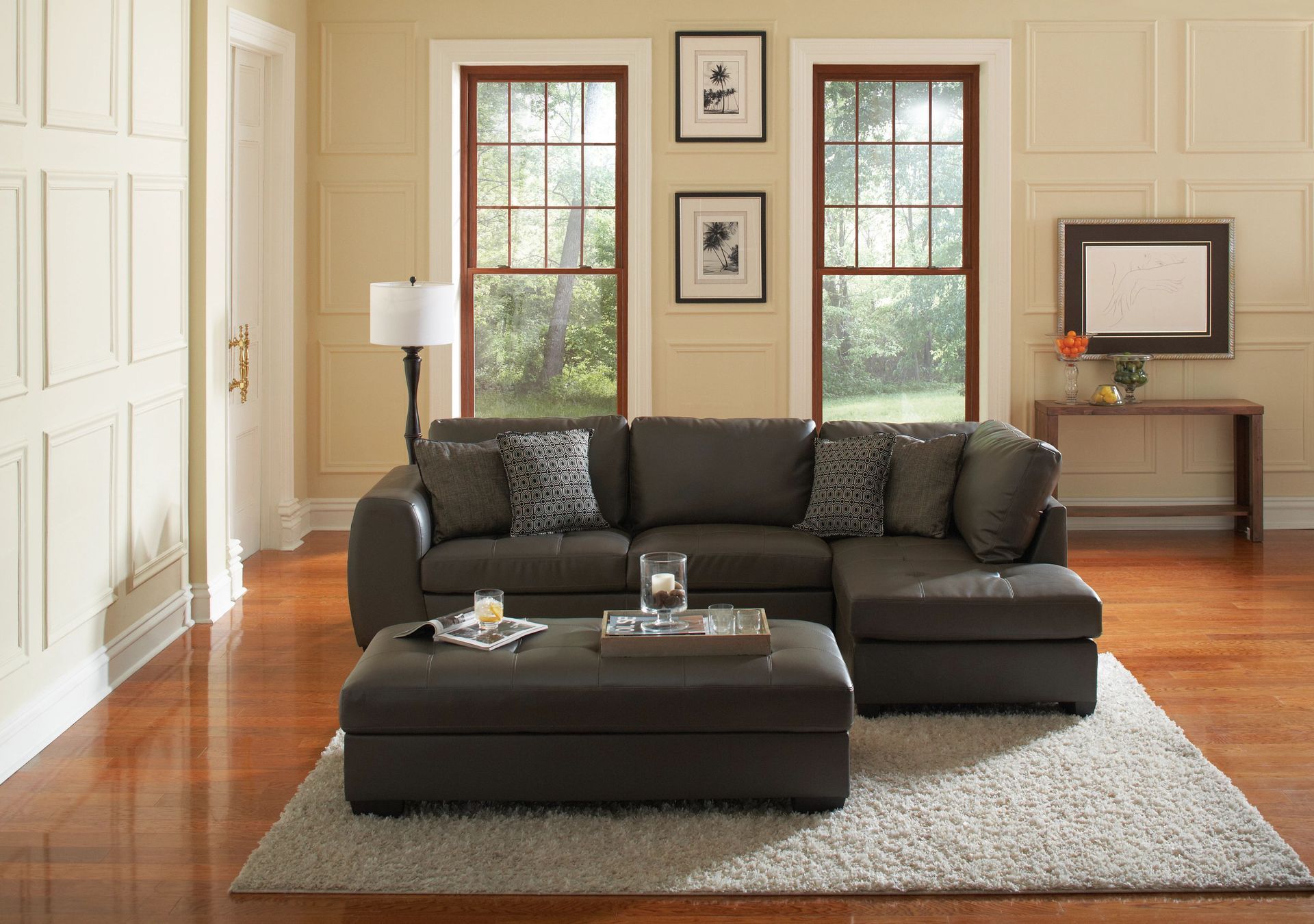 A dark brown sectional sofa with a matching ottoman on a light rug in a room with paneled cream walls and two windows.