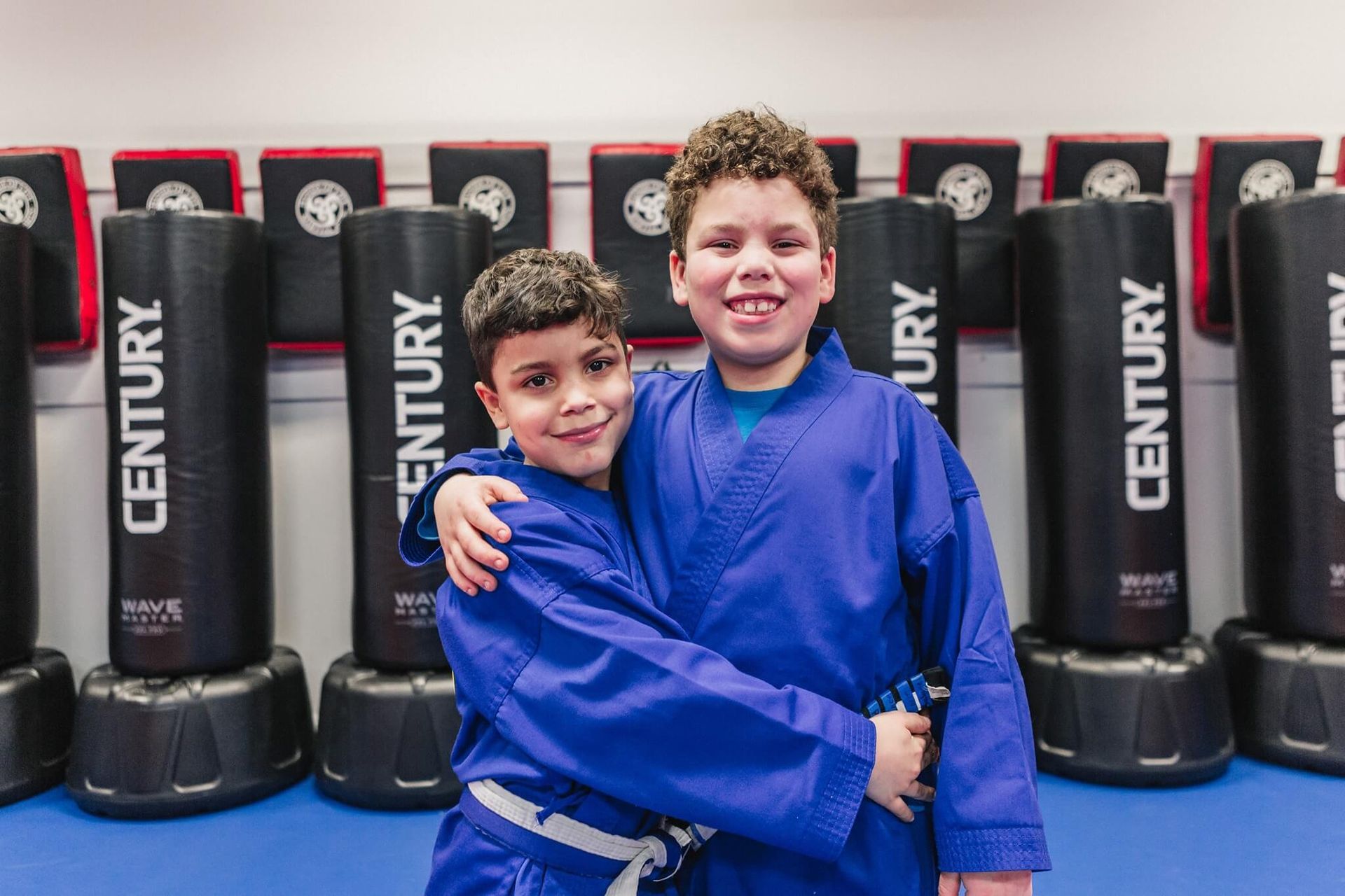 Two young boys in blue karate uniforms are hugging each other in a gym.