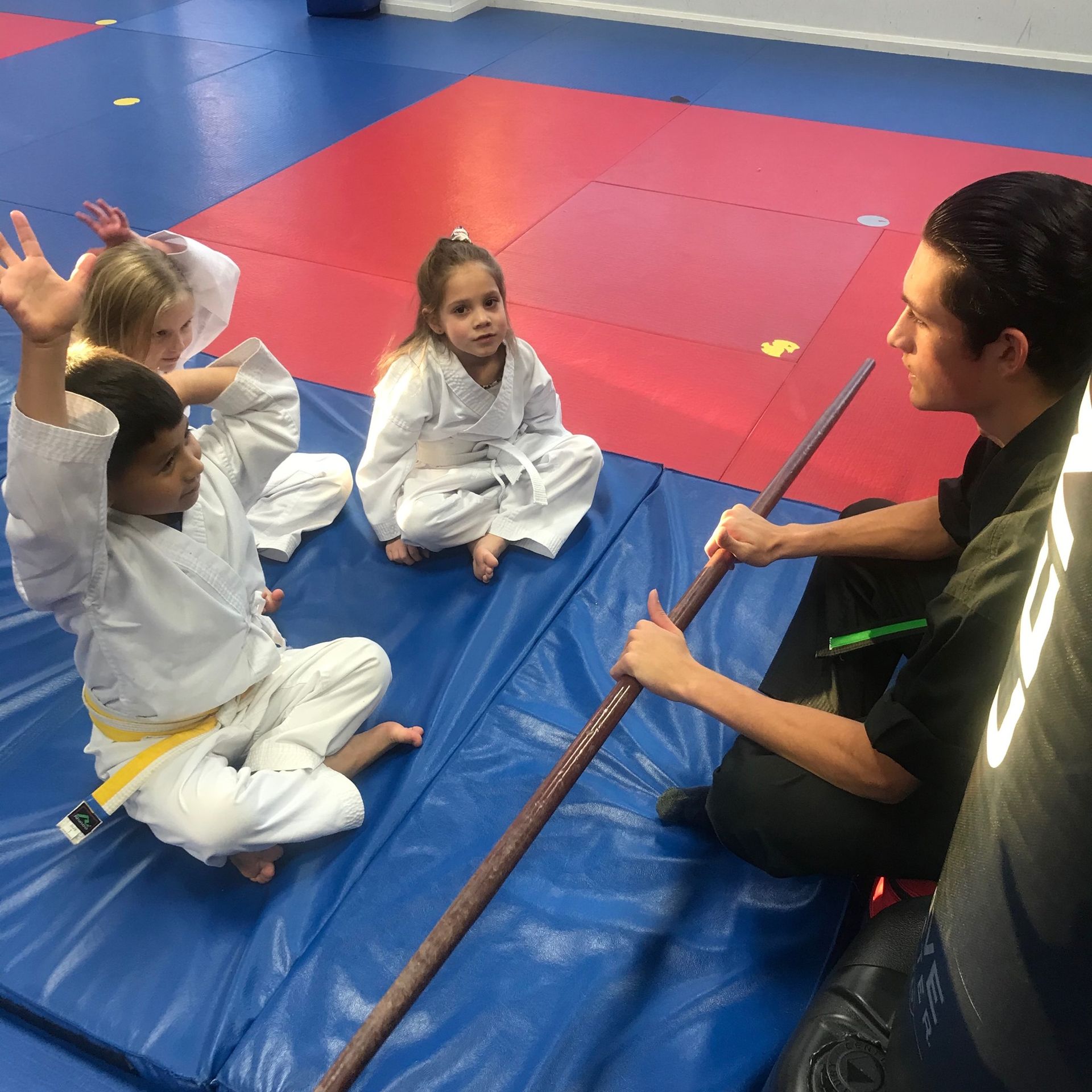 A group of children are sitting on a mat with a man holding a stick.