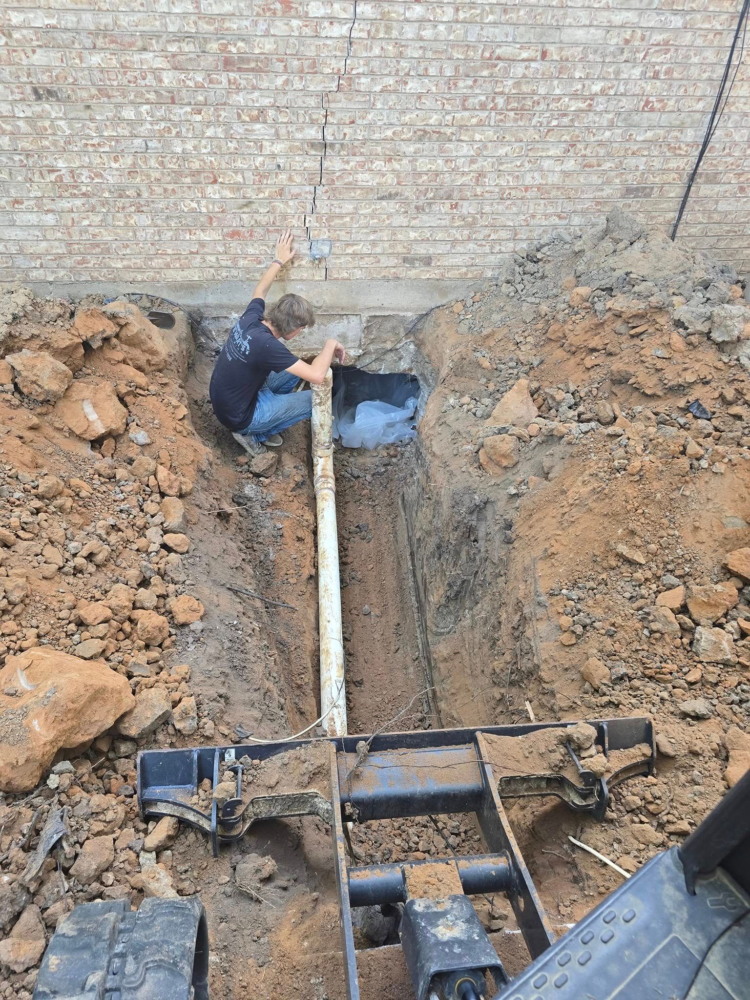 Person in trench works on white pipe near brick wall.