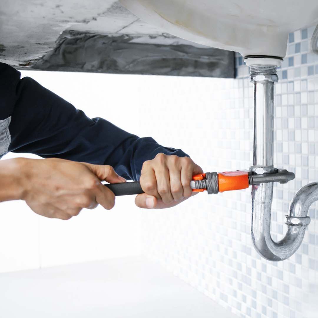 Plumber using a wrench to work on a sink's pipes; hands visible, tiles in background.