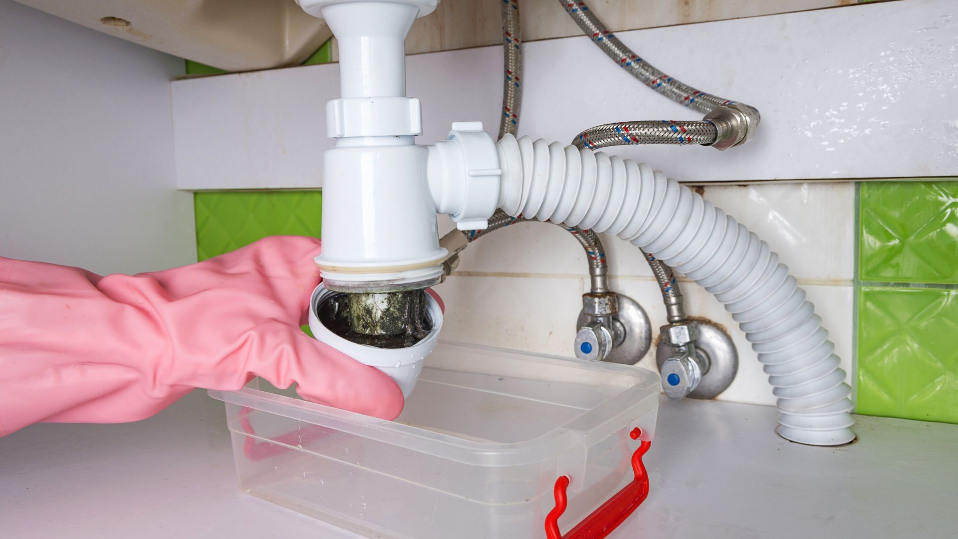 A gloved hand holds a sink trap above a container.