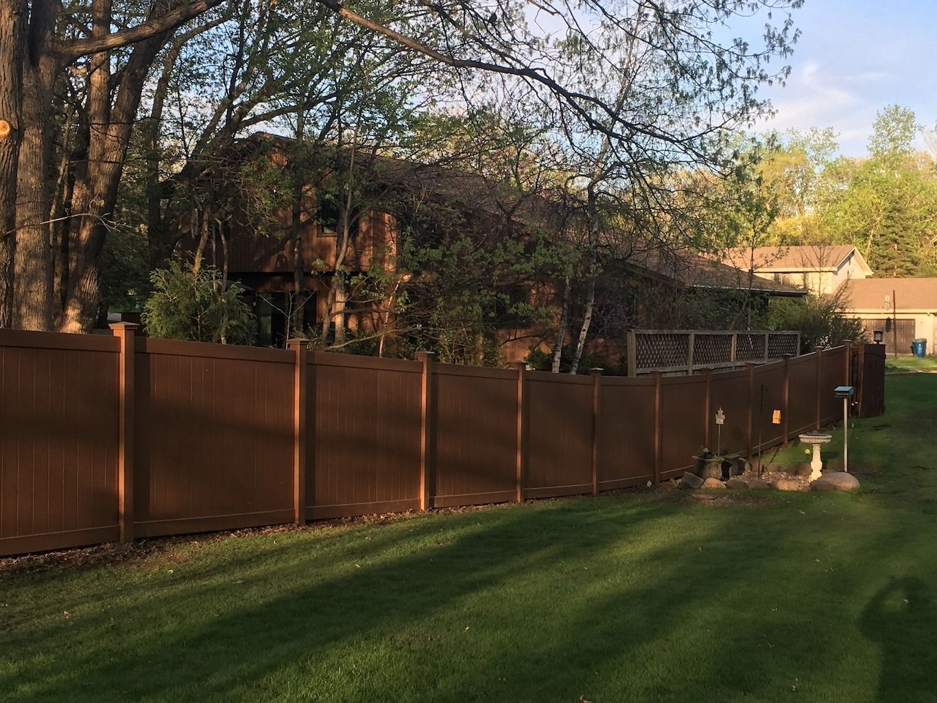Brown fence along a grassy backyard with trees and a house in the background