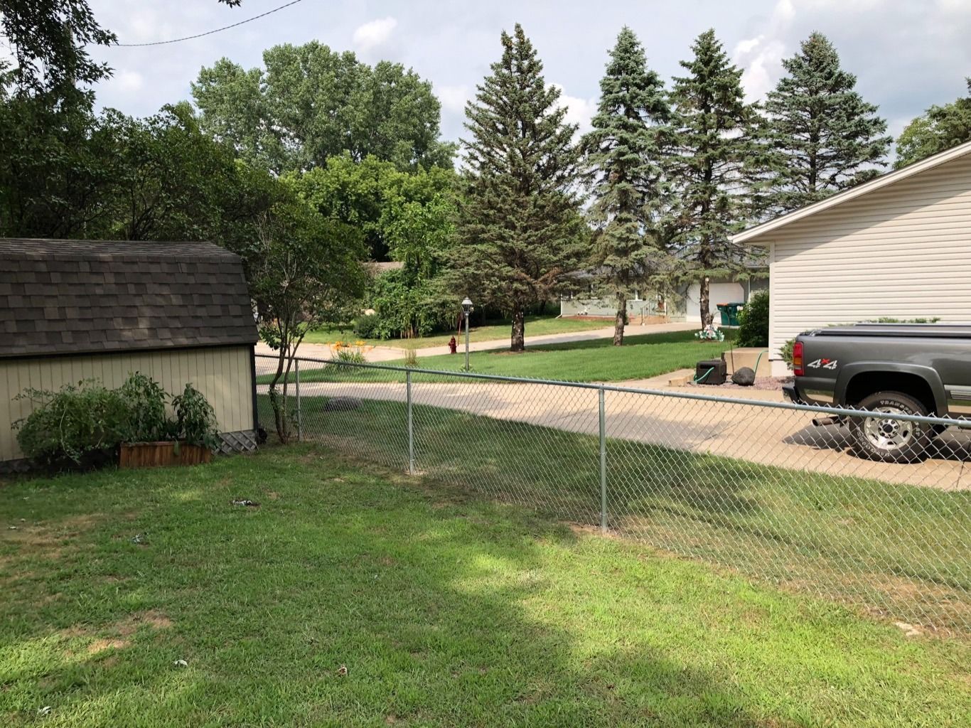 Gravel driveway between a shed and a house, with a pickup truck and green yard under trees