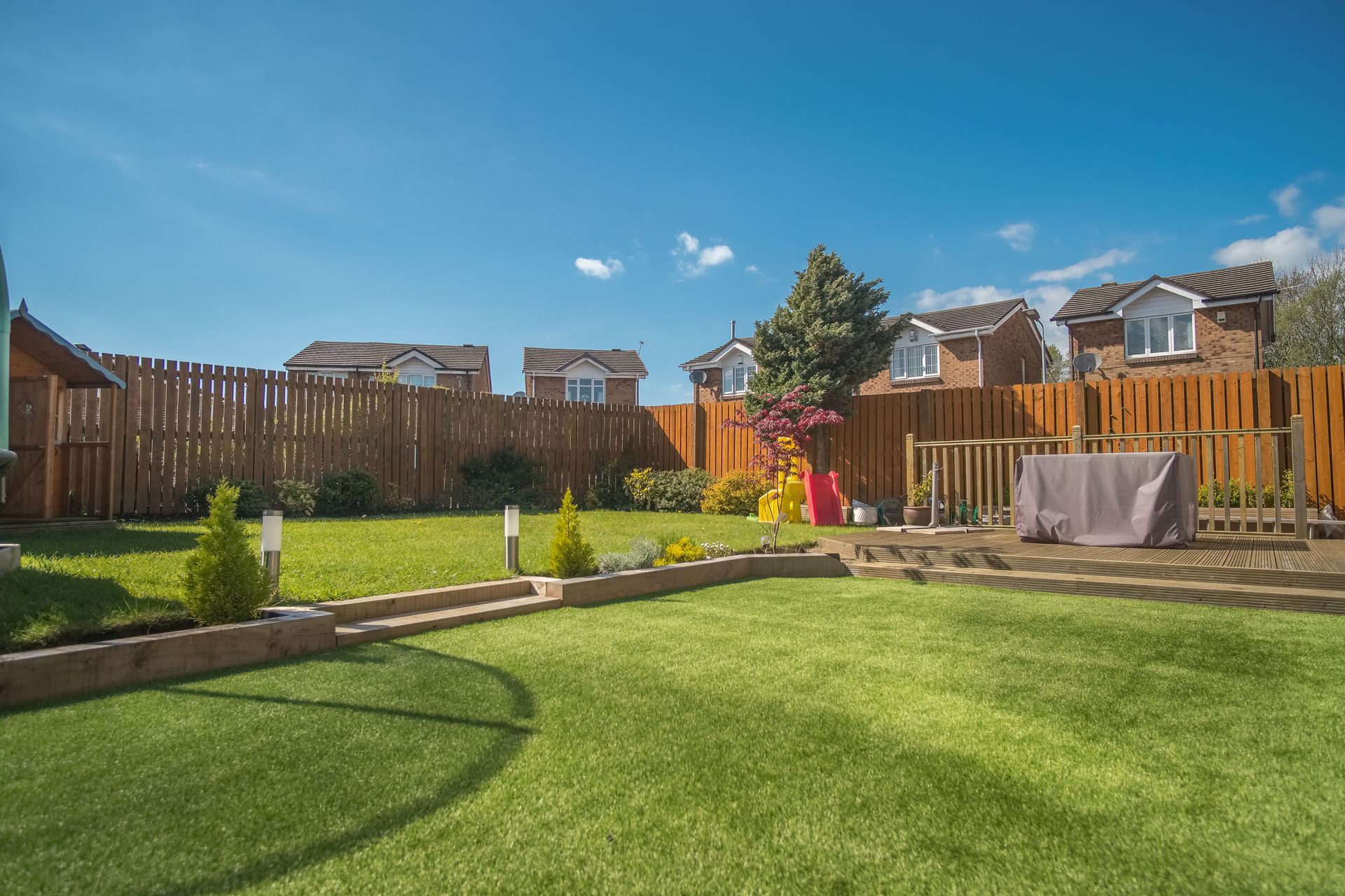 Sunny backyard with a green lawn, flower beds, wooden fence, and patio furniture under a blue sky.