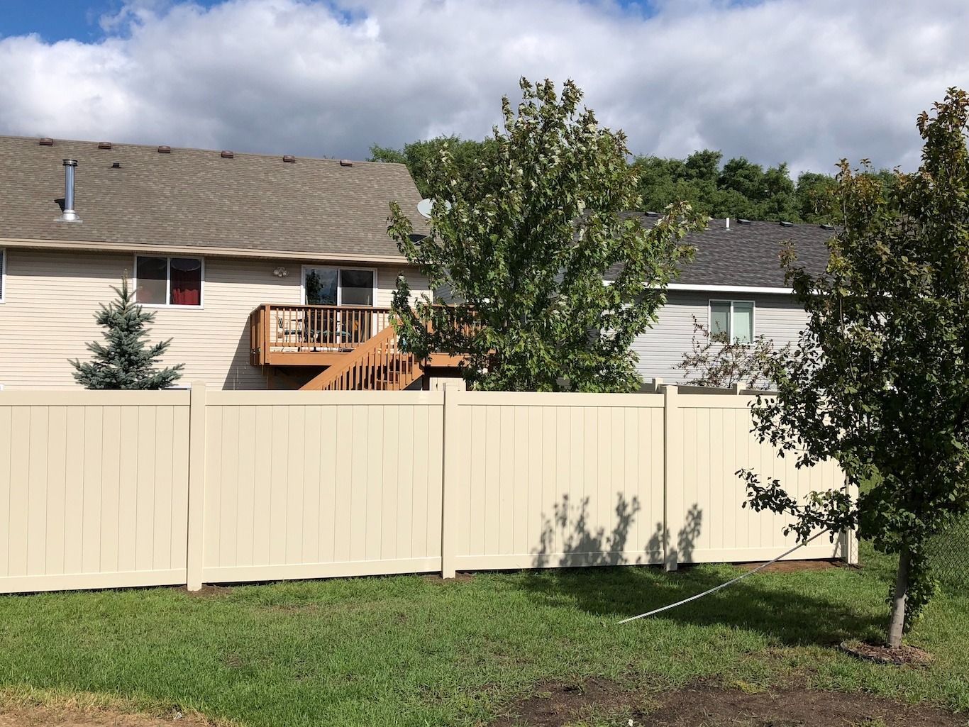Suburban backyard with beige fence, green lawn, trees, and a house with a wooden deck under cloudy sky