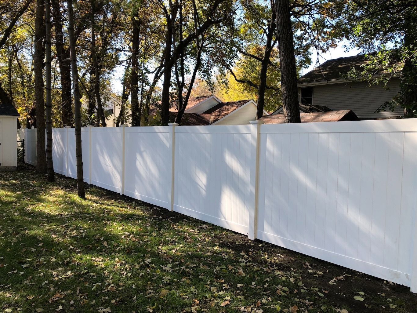 White fence along a grassy yard with trees casting shadows, and houses visible behind it.