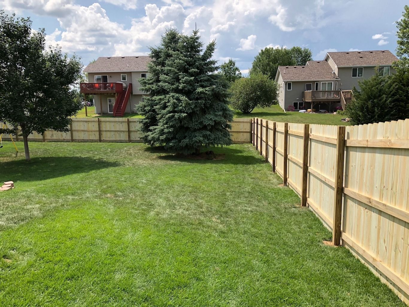 Backyard with green lawn, wooden fence, trees, and houses under a partly cloudy sky