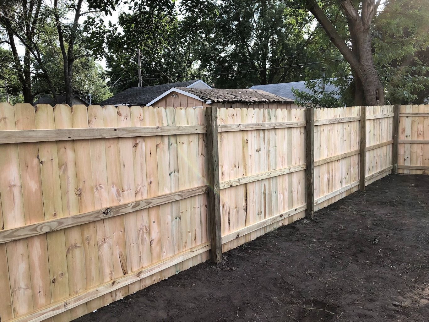 New wooden privacy fence along a dirt yard with trees and a neighboring roof in the background