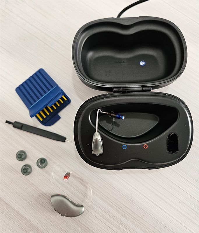 Open black earbud case with accessories: blue battery, cables, screws, and a small gray part on a light surface