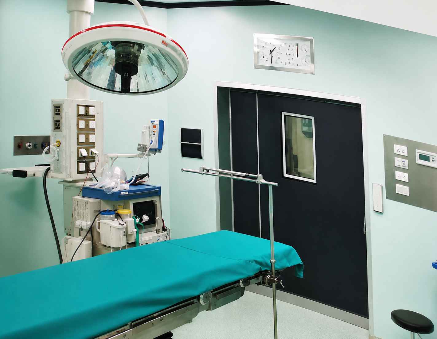 Sala operatoria vuota con tavolo chirurgico color verde acqua, lampada a soffitto e porte nere chiuse.
