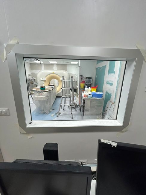 Sala di diagnostica per immagini vista attraverso una finestra, con un grande scanner, apparecchiature e un monitor di computer in primo piano.