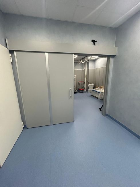 Stretto corridoio d'ospedale con pavimento azzurro, pareti grigie e porta scorrevole che si apre su una sala per trattamenti.