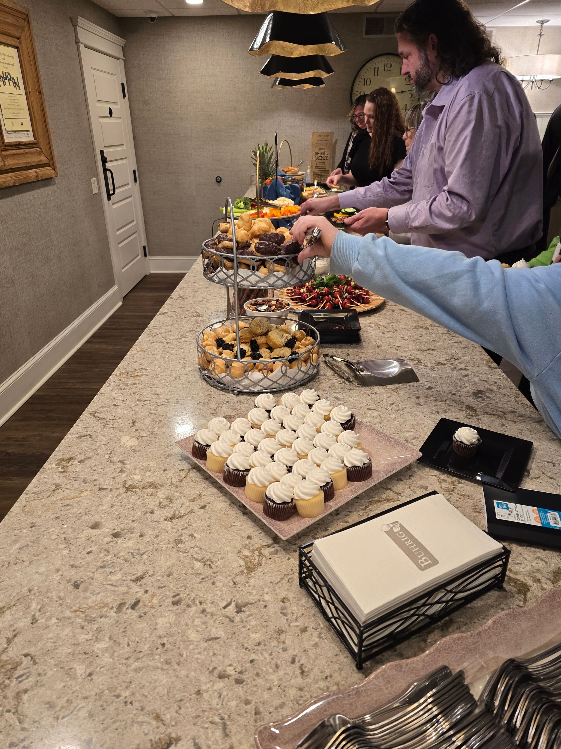 Dessert buffet on a counter with cupcakes, pastries, fruit, and people serving themselves